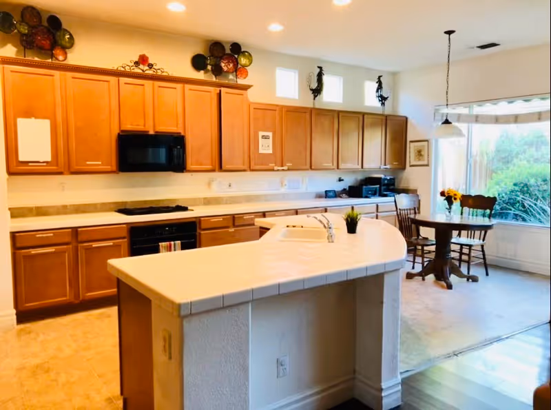 A bright kitchen with wooden cabinets, a white tiled countertop island with a sink, and a small round dining table with two chairs near a large window with a view of greenery outside. Decorative plates and rooster figurines are displayed above the cabinets.