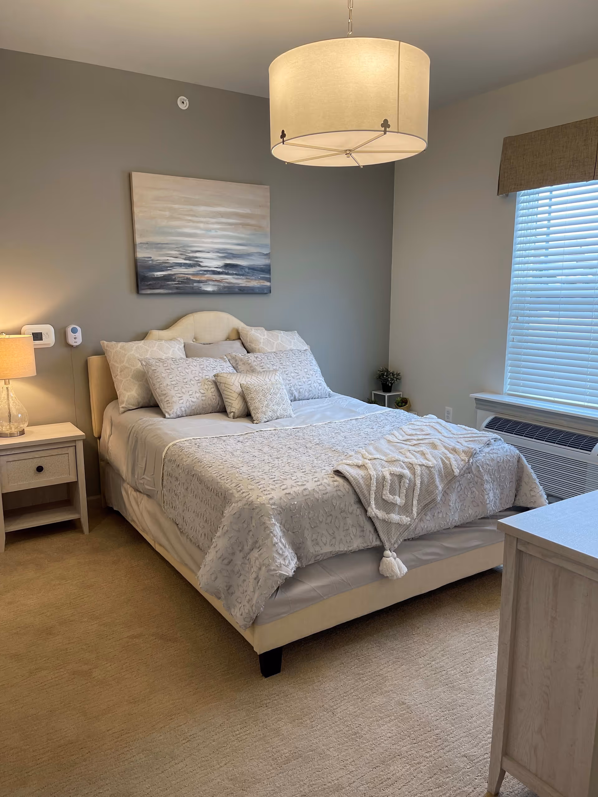 A neatly made bed with multiple pillows and a textured blanket in a softly lit bedroom. The room features a nightstand with a lamp, a window with blinds and a valance, a small plant on a side table, a large ceiling light fixture, and a piece of abstract art hanging above the bed.