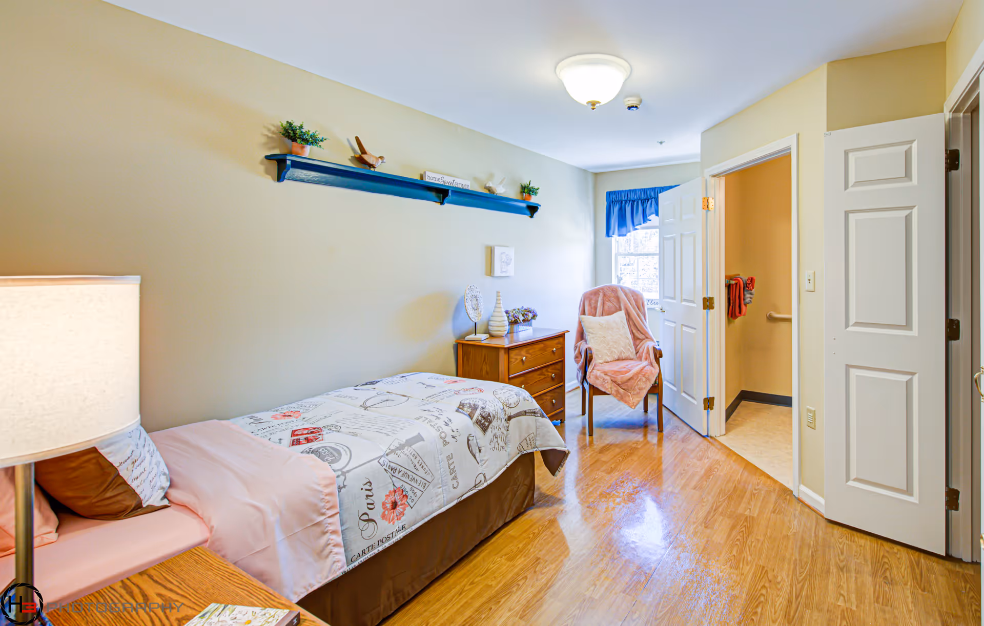 Small furnished bedroom with a twin bed, wooden dresser, armchair, and an open doorway to a bathroom.
