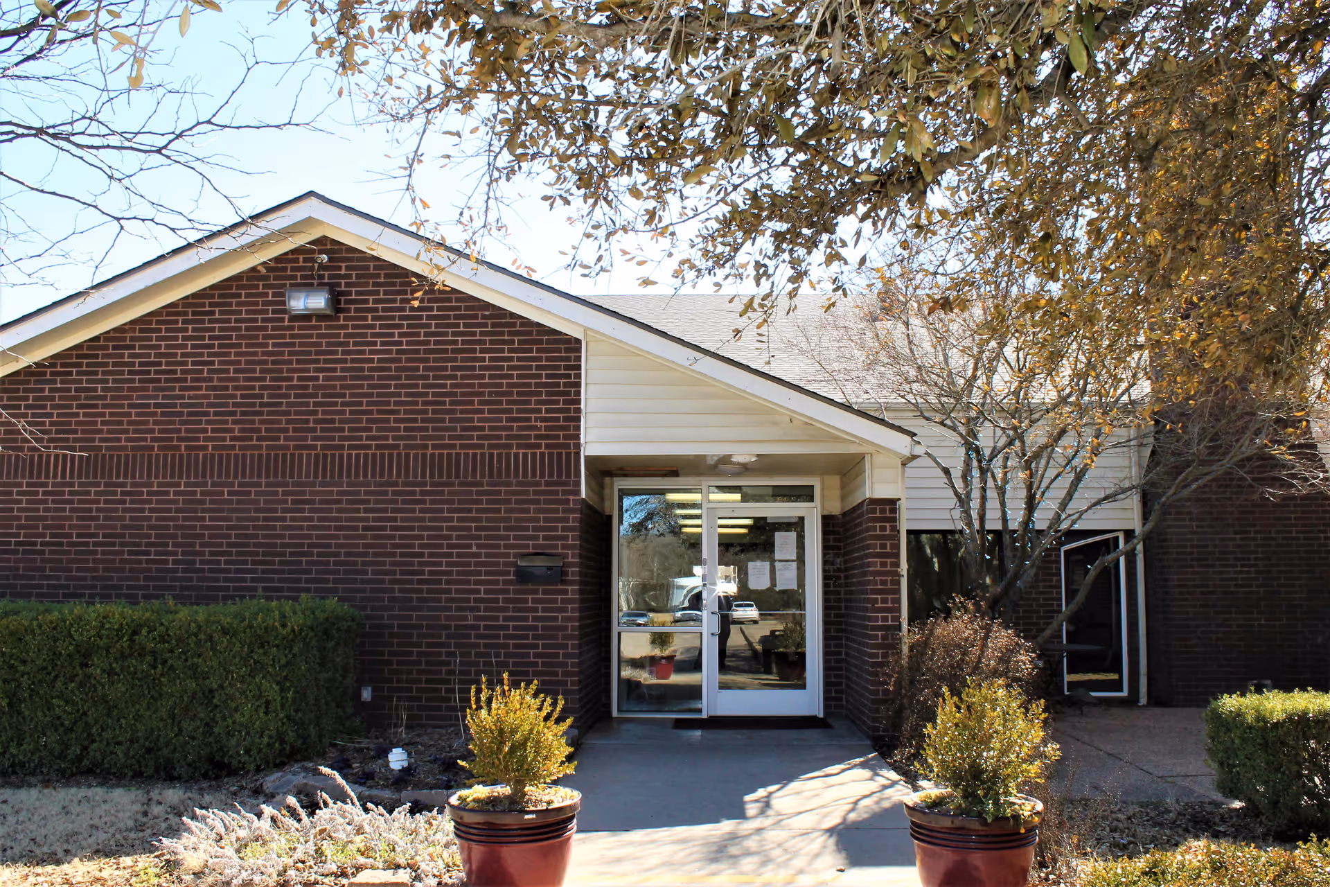Exterior view of a single-story brick building with a peaked roof and a glass entrance door. There are two potted plants on either side of the walkway leading to the entrance, bushes along the building, and leafless trees overhead.