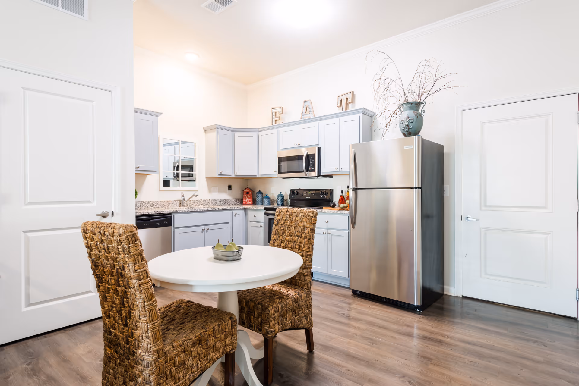 A bright kitchen area with light gray cabinets, a stainless steel refrigerator, microwave, and stove. There is a small round white dining table with two woven chairs in the foreground. Decorative items including a vase with branches and letters spelling 'EAT' are placed on top of the cabinets.