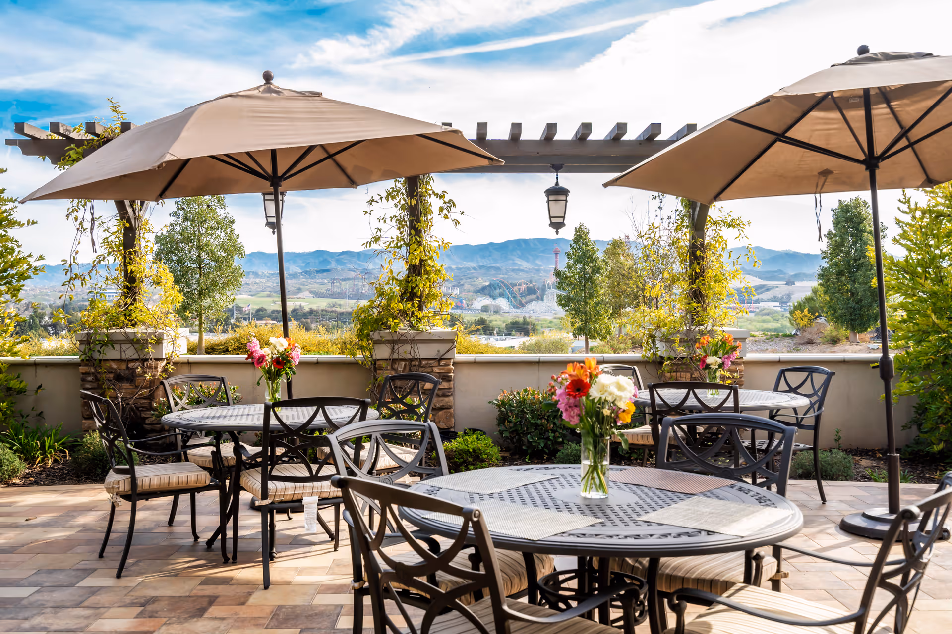 Outdoor patio area with round metal tables and cushioned chairs under large beige umbrellas. Each table has a vase with colorful flowers. The patio overlooks a scenic view of trees, hills, and a partly cloudy sky.