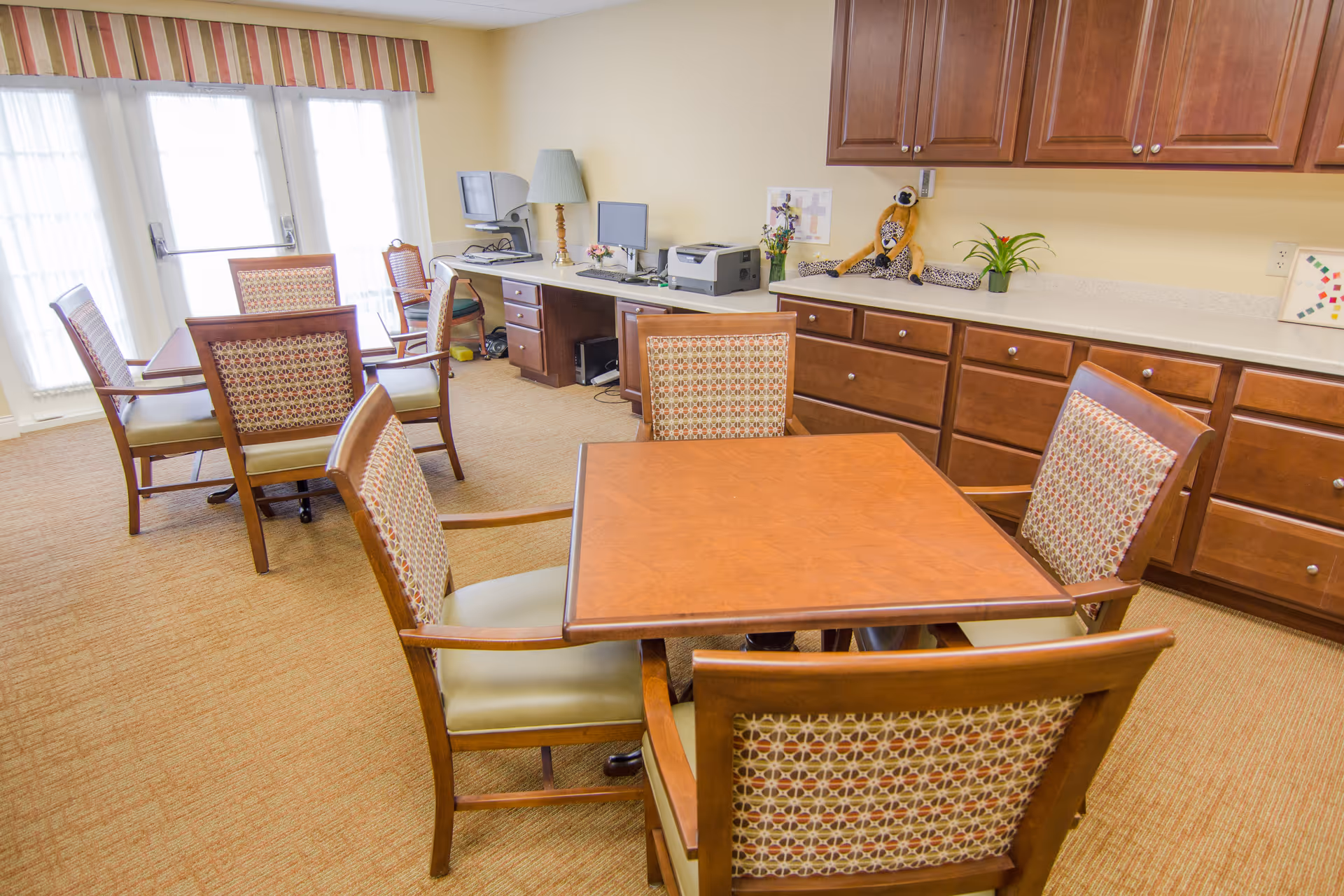 A well-lit room with multiple wooden tables and chairs arranged for seating. The chairs have patterned upholstery and light green cushions. Along one wall, there are wooden cabinets and a countertop with decorative items including a stuffed animal and plants. In the background, there is a desk with a computer, printer, and a lamp. Large windows with sheer curtains and a striped valance allow natural light to fill the room.