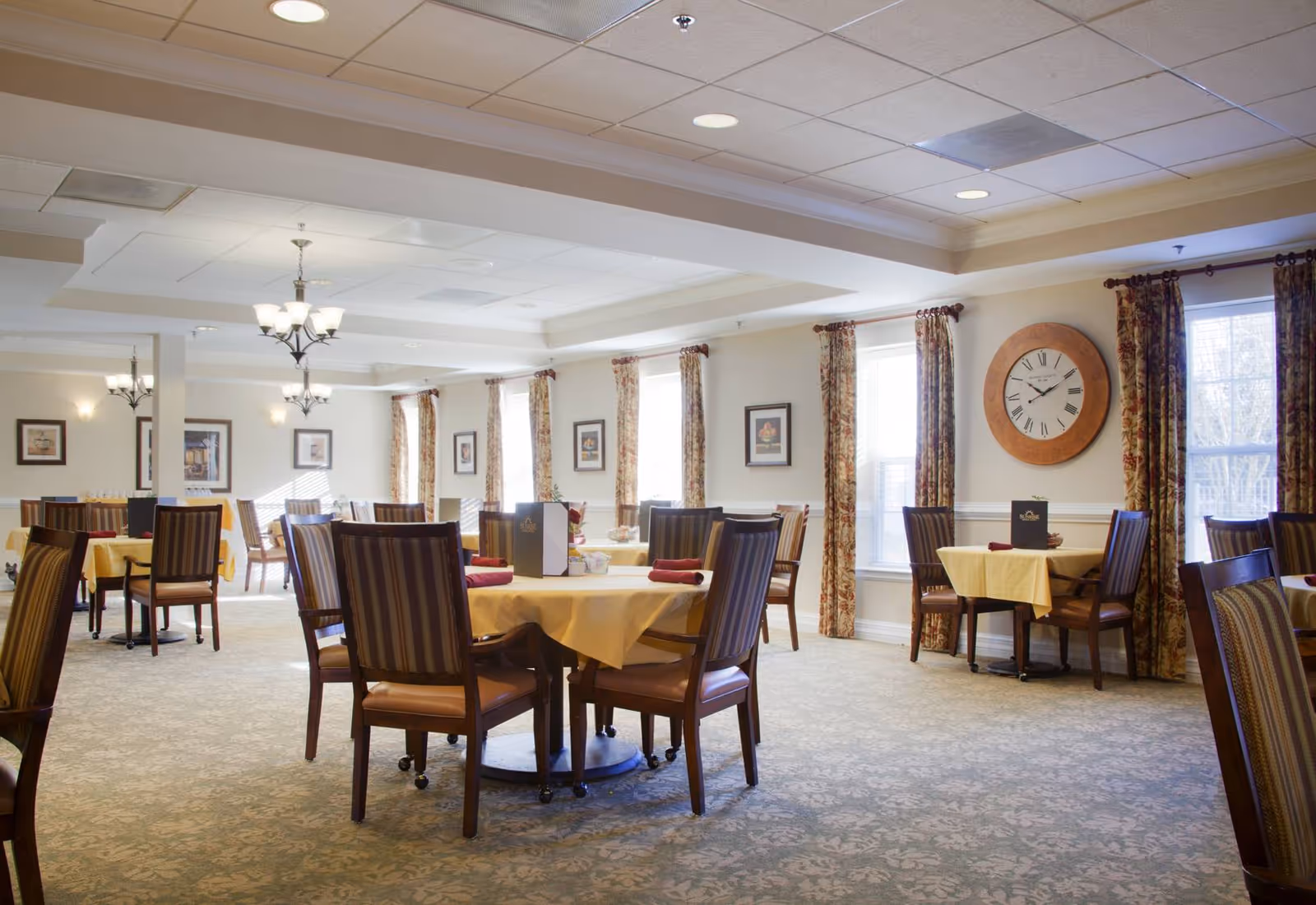 A bright dining room with multiple round tables covered with yellow tablecloths and surrounded by wooden chairs with striped cushions. The room has large windows with floral curtains, framed pictures on the walls, and a large round clock. Ceiling lights provide illumination.