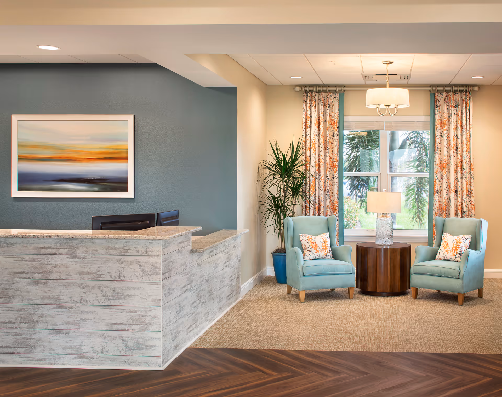 A reception area with a light wood textured front desk on the left and two teal armchairs with floral pillows on the right. Between the chairs is a round wooden table with a lamp. Behind the chairs is a window with floral curtains and a tall green plant in a blue pot. A colorful abstract painting hangs on the blue wall behind the desk.