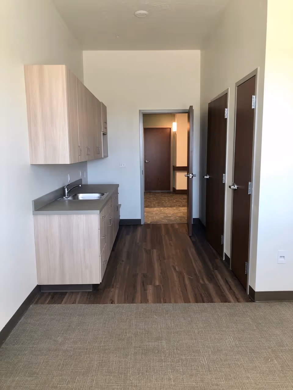 A small kitchenette area in an assisted living facility with light wood cabinets, a countertop with a sink, and a dark wood floor. There are three closed doors on the right side and an open door at the end of the room leading to a carpeted hallway.