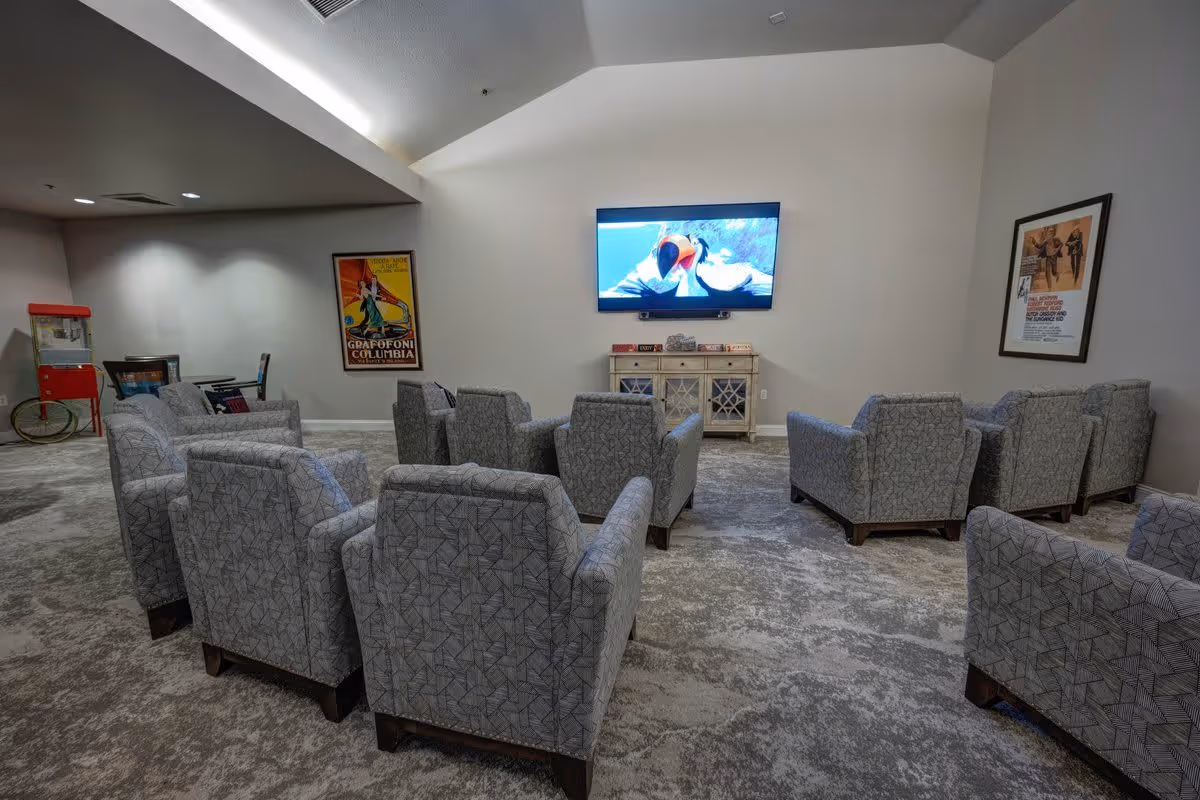 A cozy common room with multiple patterned armchairs arranged in rows facing a wall-mounted flat screen TV displaying an animated movie. The room has a light gray carpet and neutral-colored walls with two framed vintage posters. A small cabinet sits beneath the TV, and a red popcorn machine is visible in the corner near a small table and chairs.
