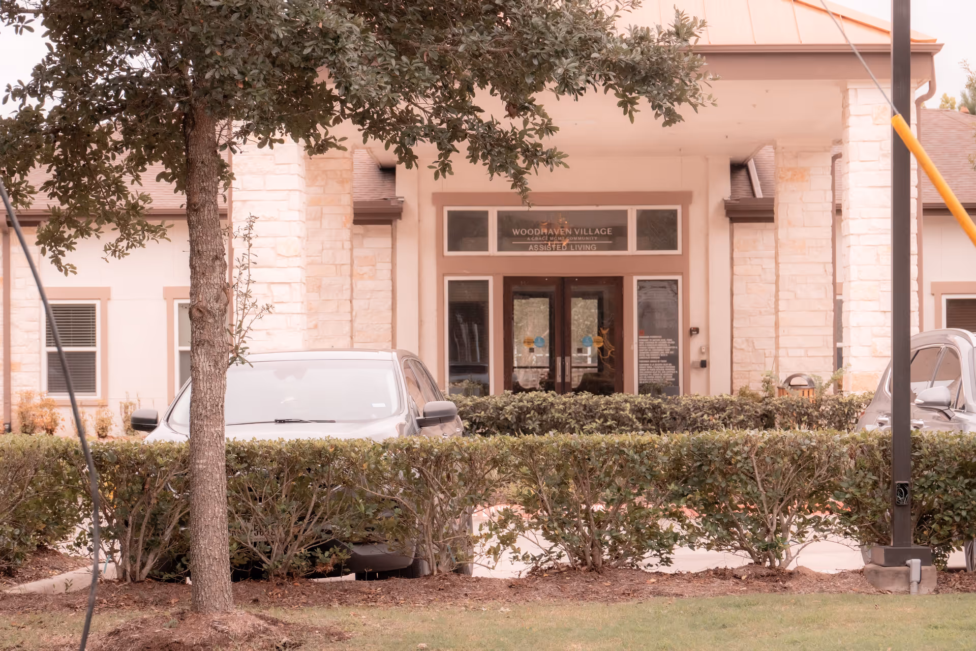Front entrance of Woodhaven Village assisted living facility with stone pillars, glass double doors, and a sign above the entrance. There are bushes and parked cars in front of the building, and a tree partially obscures the view.