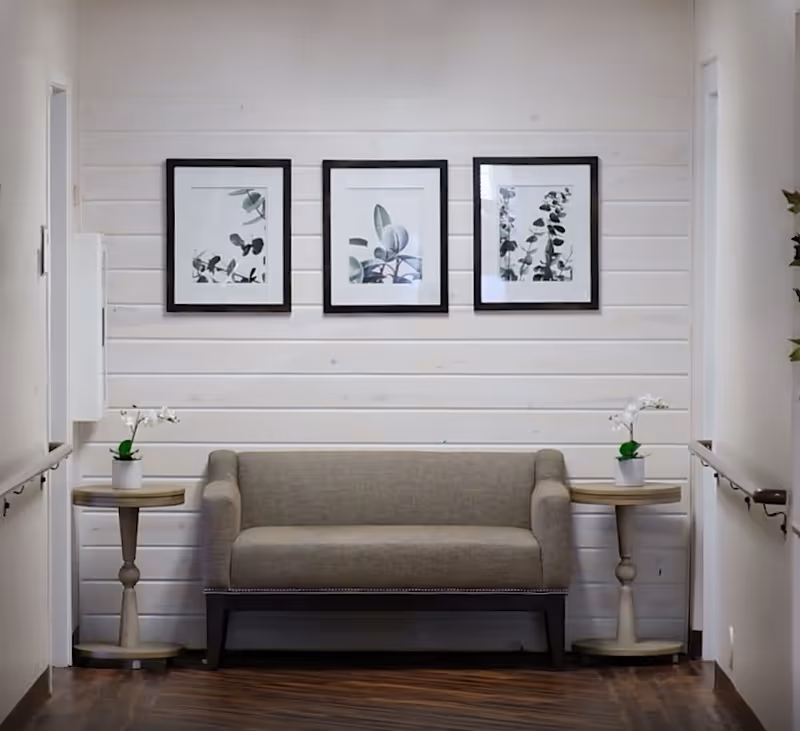 A small seating area with a gray upholstered bench between two round wooden side tables, each holding a small white orchid plant. Behind the bench is a white shiplap wall with three framed botanical prints hanging above. The floor is dark wood, and there are handrails on both sides of the hallway.