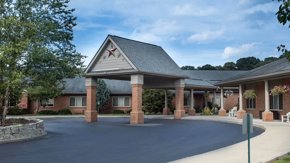 Exterior view of Bickford of Lancaster facility showing a covered entrance with brick pillars and a triangular roof decorated with stars. The building is single-story with brick walls and several windows. There are trees, hanging flower baskets, and a curved driveway in front of the entrance under a partly cloudy sky.