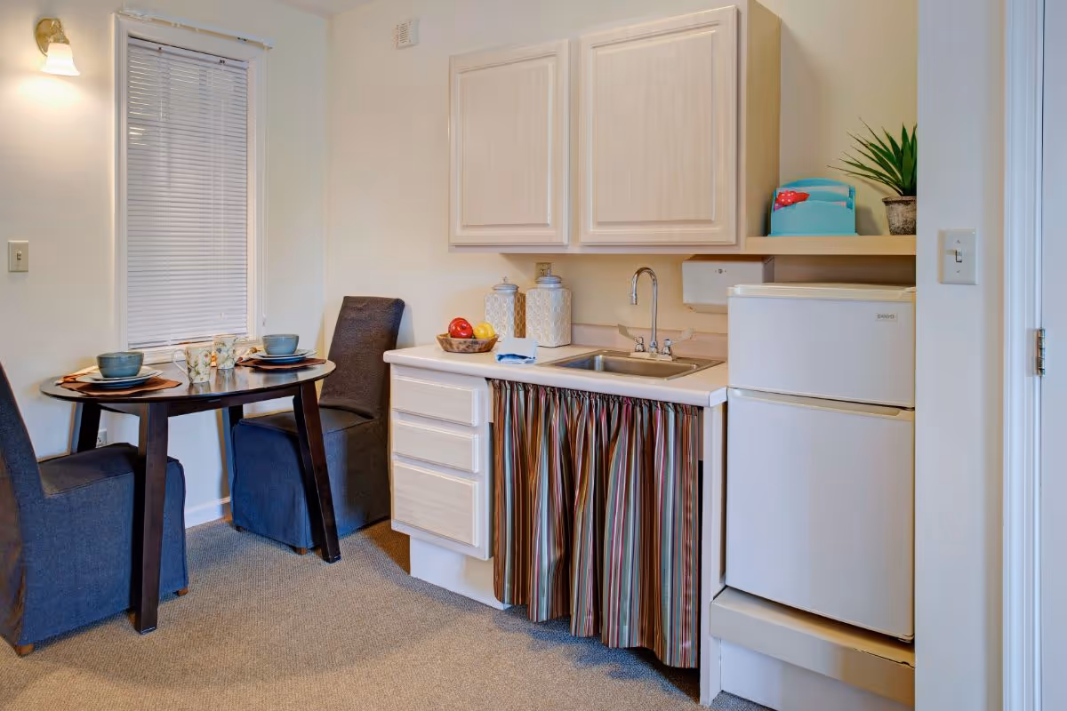 Small kitchenette with a sink, mini refrigerator, upper cabinets and a round dining table set with two chairs.