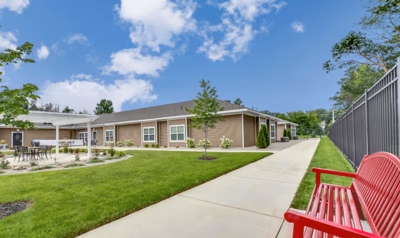 Outdoor view of Candlestone Assisted Living & Memory Care facility showing a single-story building with beige siding and white trim. There is a concrete walkway bordered by green grass, a red bench on the right side, and a patio area with tables and chairs under a white pergola. Trees and shrubs are planted around the building and along the walkway under a partly cloudy blue sky.