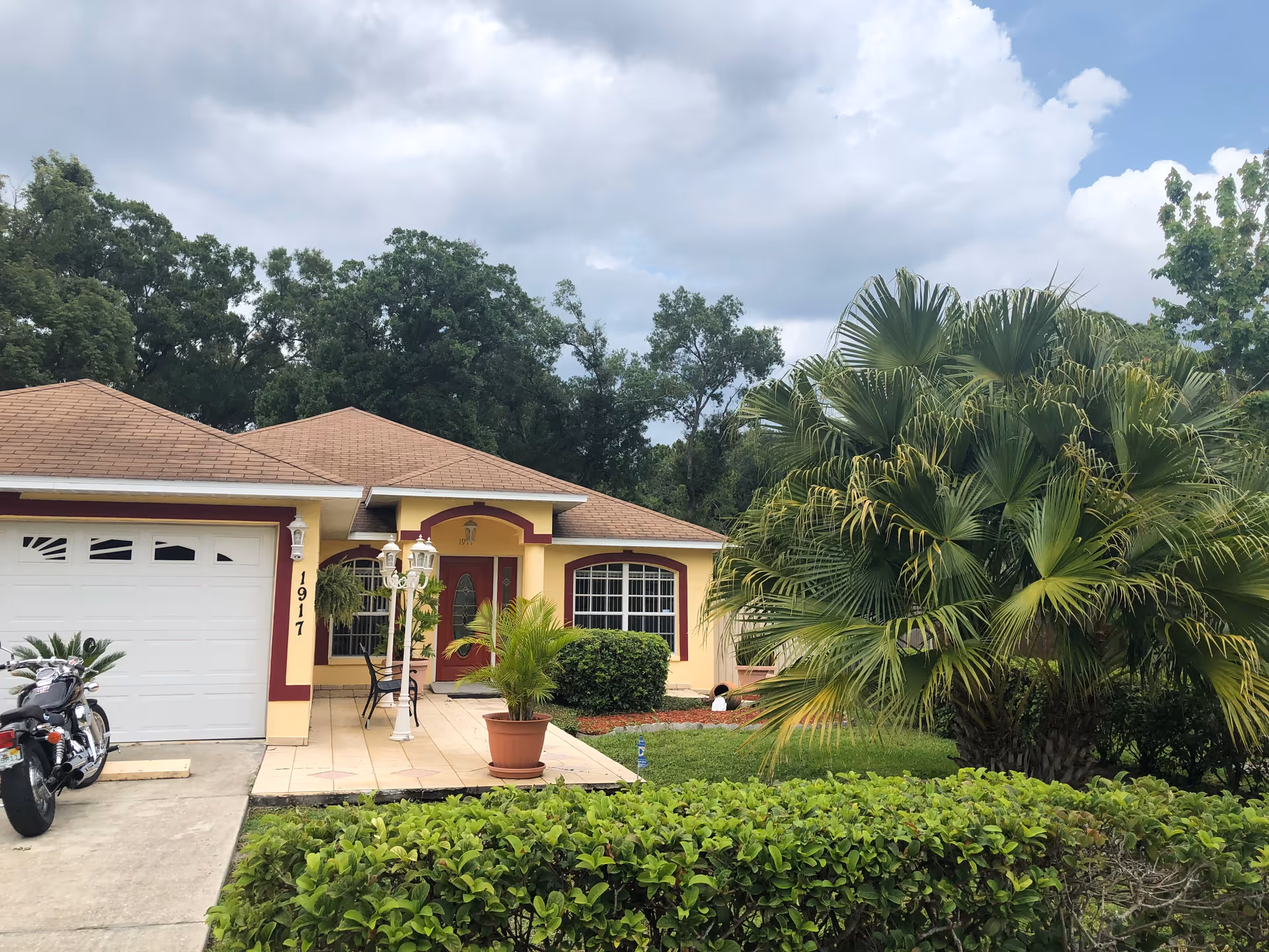 Front exterior of a single-story yellow house with a garage, tiled porch, driveway and a large palm tree in the front yard.