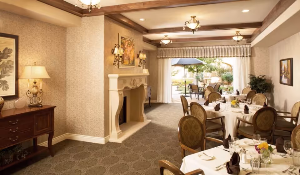 A warmly lit dining room with round tables covered in white tablecloths, set with glasses, napkins, and flowers. The room features a decorative fireplace, wall sconces, framed artwork, and a side table with a lamp. Large windows with curtains open to an outdoor patio area with tables, chairs, and an umbrella.