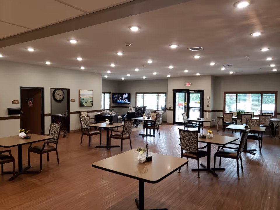 A spacious dining room in a senior living facility with multiple square tables and patterned chairs arranged neatly. The room has wooden flooring, bright ceiling lights, large windows letting in natural light, and a TV mounted on the far wall. A person in a wheelchair is visible near the TV, and there are small flower arrangements on the tables.
