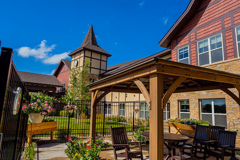 Outdoor patio area at Guardian Angels Engel Haus Senior Living - Albertville featuring a wooden pergola with seating, surrounded by flowers and greenery, with the senior living building in the background under a clear blue sky.