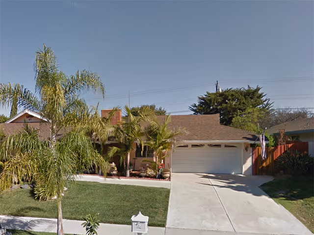 Single-story residential building with a brown shingle roof, a two-car garage, and a driveway. The front yard has green grass, several palm trees, and other plants. A white mailbox is visible near the sidewalk. The sky is clear and blue.