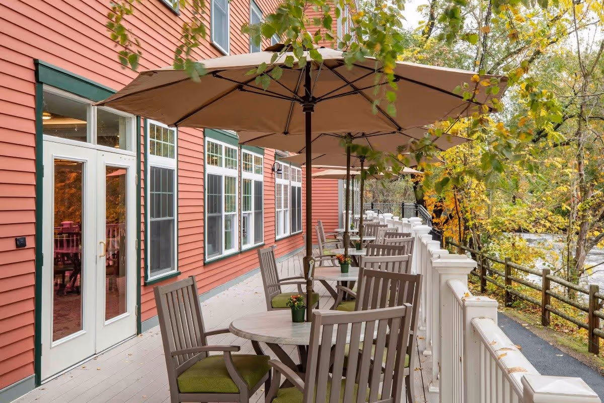 Outdoor patio area at The Falls at Cordingly Dam with round tables, wooden chairs with green cushions, large beige umbrellas, and a white railing overlooking a wooded area with a river.