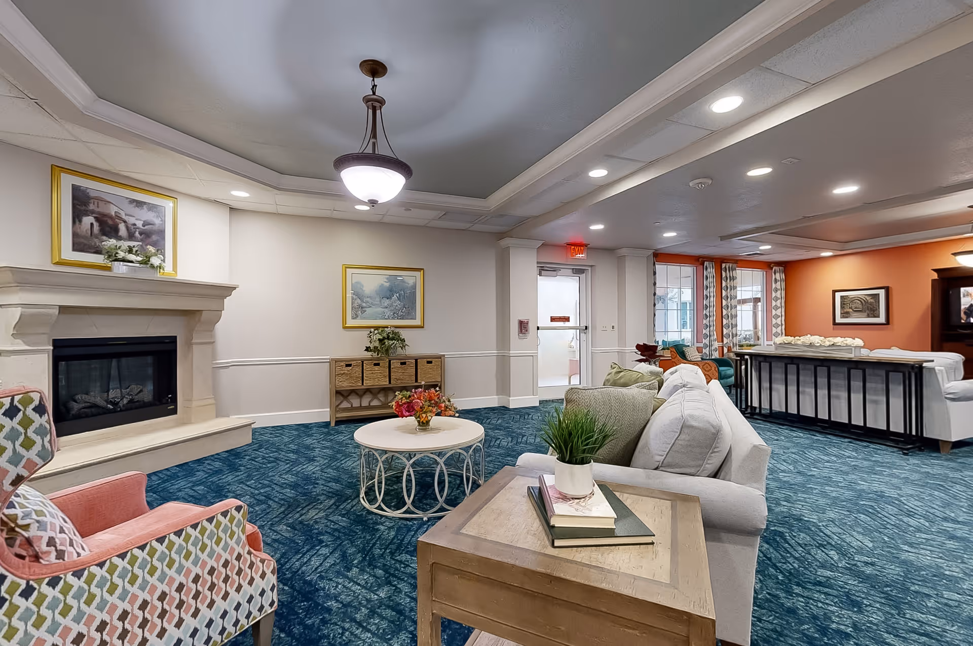 A cozy living room area in a senior living facility featuring a fireplace with a painting above it, a patterned armchair, a round coffee table with a flower arrangement, a side table with books and a potted plant, and several sofas. The room has blue carpet, light-colored walls with framed artwork, and a ceiling light fixture. Large windows with patterned curtains and an orange accent wall are visible in the background.