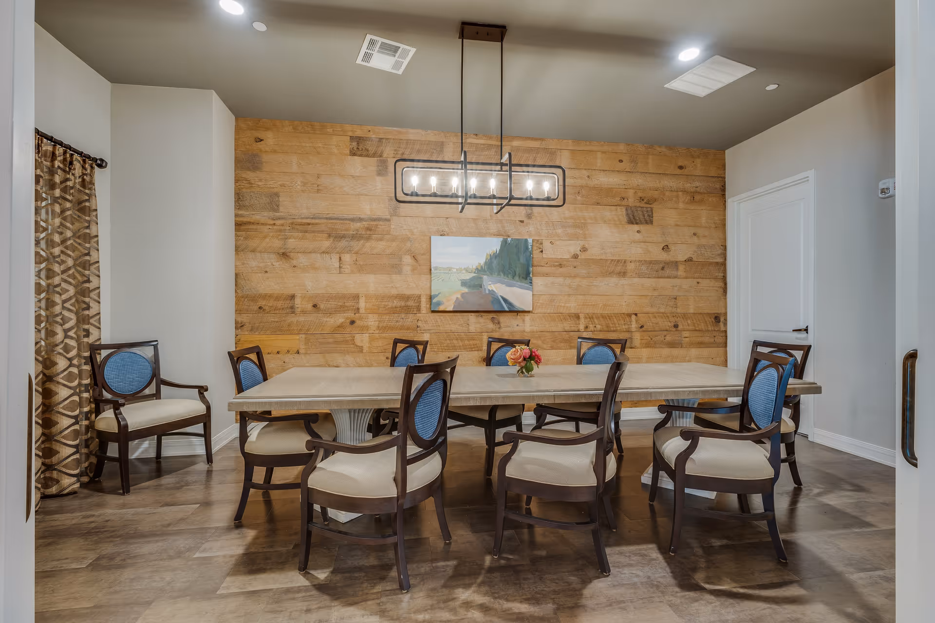 A dining room with a rectangular table surrounded by eight wooden chairs with blue upholstered backs and cream-colored seats. The room features a wooden plank accent wall with a landscape painting hanging in the center. A modern rectangular chandelier with exposed bulbs hangs above the table. The floor is wood, and there is a patterned curtain on the left side of the room.