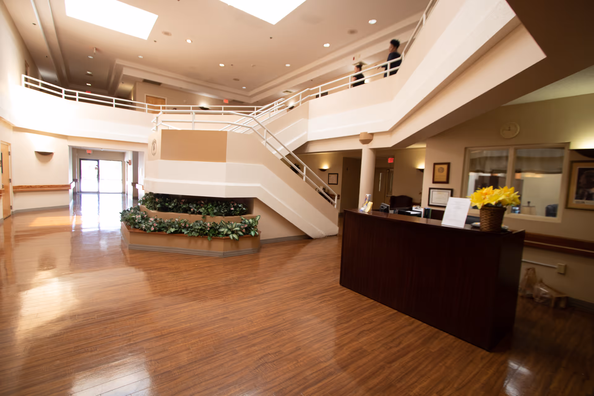 Interior view of a nursing and rehabilitation center lobby with a wooden reception desk adorned with a basket of yellow flowers. The space features polished wooden floors, a staircase with white railings leading to an upper level, and a planter with green plants at the base of the stairs. There are doors and windows along the walls, and two people are visible walking on the upper level.
