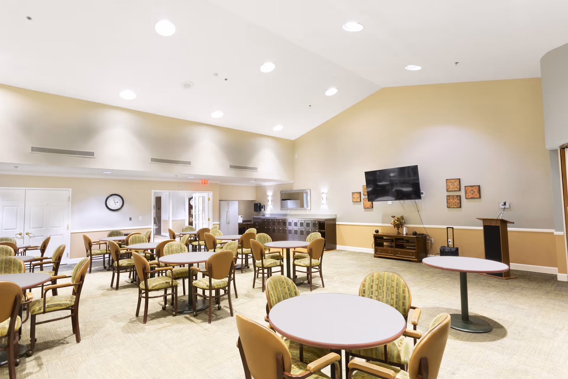 Bright communal dining/activity room with multiple round tables and chairs, a wall-mounted TV, and cabinetry.