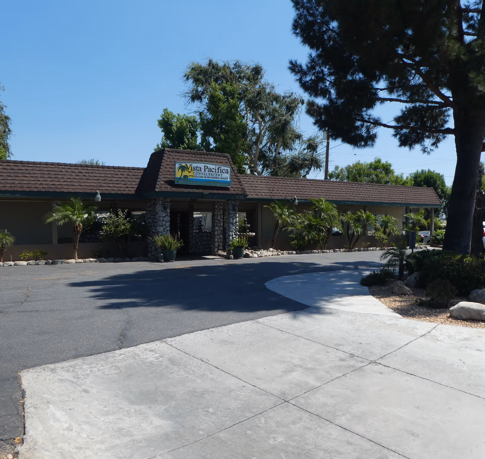 Exterior view of Vista Pacifica Convalescent facility showing a single-story building with a stone entrance, surrounded by palm trees and other greenery under a clear blue sky.