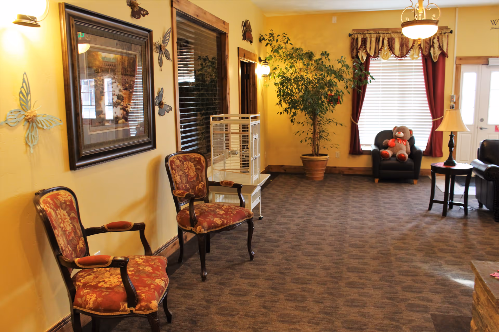 Interior lounge with upholstered chairs, a potted plant, window with curtains, and a teddy bear on an armchair.