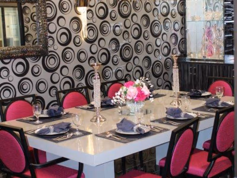 A dining room with a rectangular white table set for eight people. The chairs have black frames and bright pink cushions. The table is set with black placemats, folded dark napkins, glassware, and silverware. Two tall silver candle holders and a floral centerpiece with pink and white flowers are on the table. The wall behind features a black and white circular pattern wallpaper, a large ornate mirror, and a glass display cabinet.