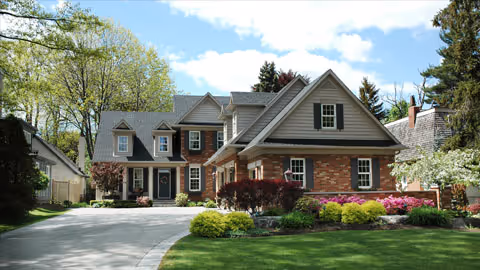 Large two-story brick and siding house with a curved driveway, manicured lawn, shrubs, and trees under a partly cloudy sky.