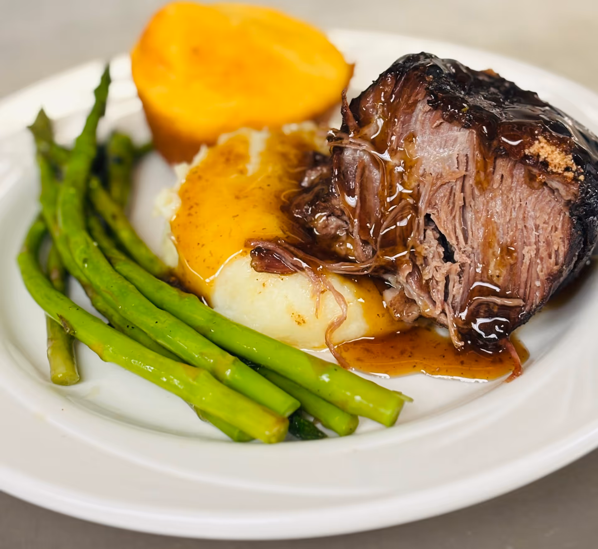 A plate of food featuring a serving of asparagus, mashed potatoes topped with brown gravy, a piece of roasted meat with gravy, and a cornbread muffin.