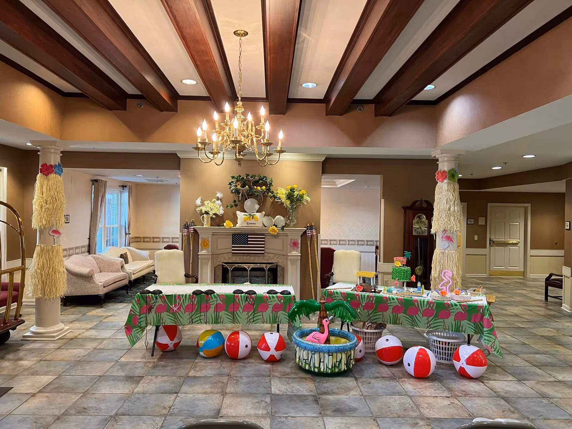 A decorated assisted living common area with a flamingo-themed party table, beach balls, columns, fireplace, and chandelier.