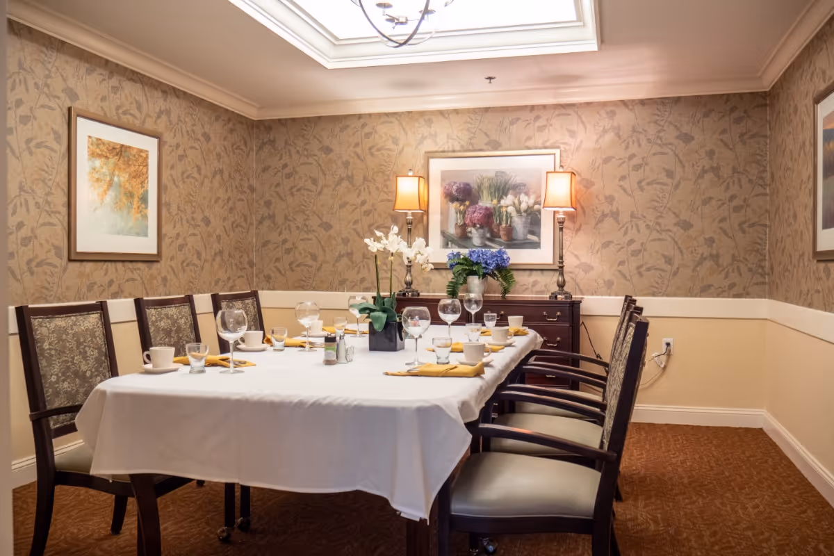A formal dining room with a rectangular table covered with a white tablecloth, set with glassware, cups, and yellow napkins. The room has patterned wallpaper, framed floral artwork on the walls, two table lamps on a sideboard, and a skylight above providing natural light.