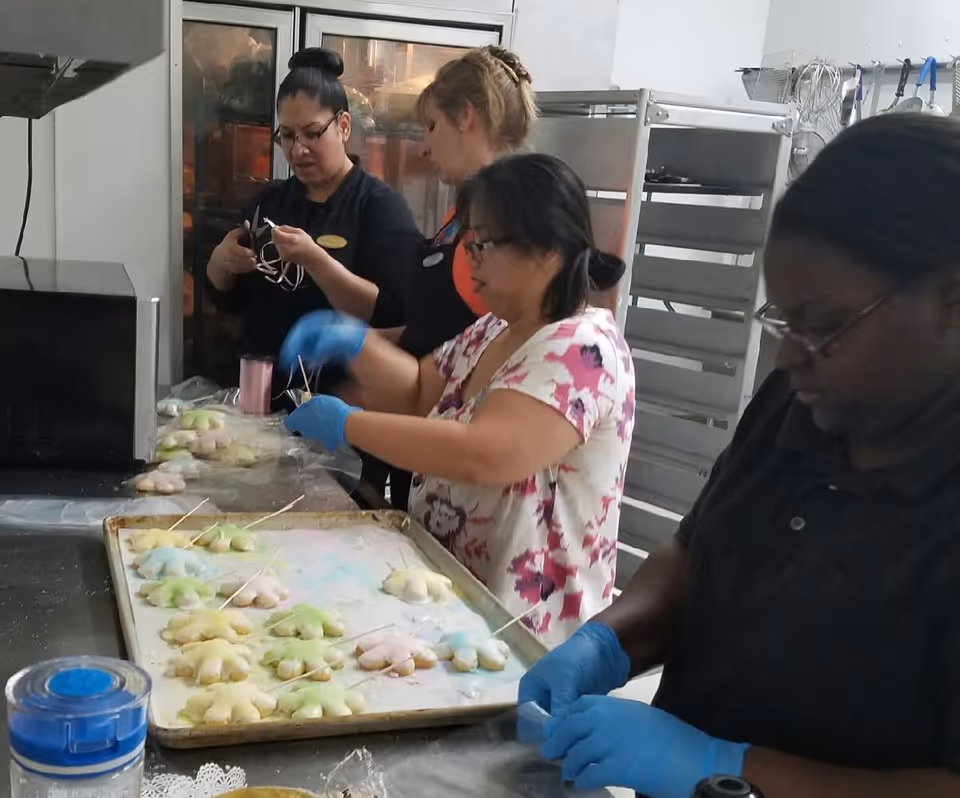 Four women working together in a kitchen preparing colorful star-shaped cookies on a baking tray. They are wearing gloves and focused on their tasks, with kitchen equipment and racks visible in the background.
