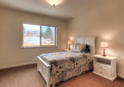 A tidy bedroom with a single bed, patterned bedspread, two bedside tables with lamps, and a window showing trees outside.
