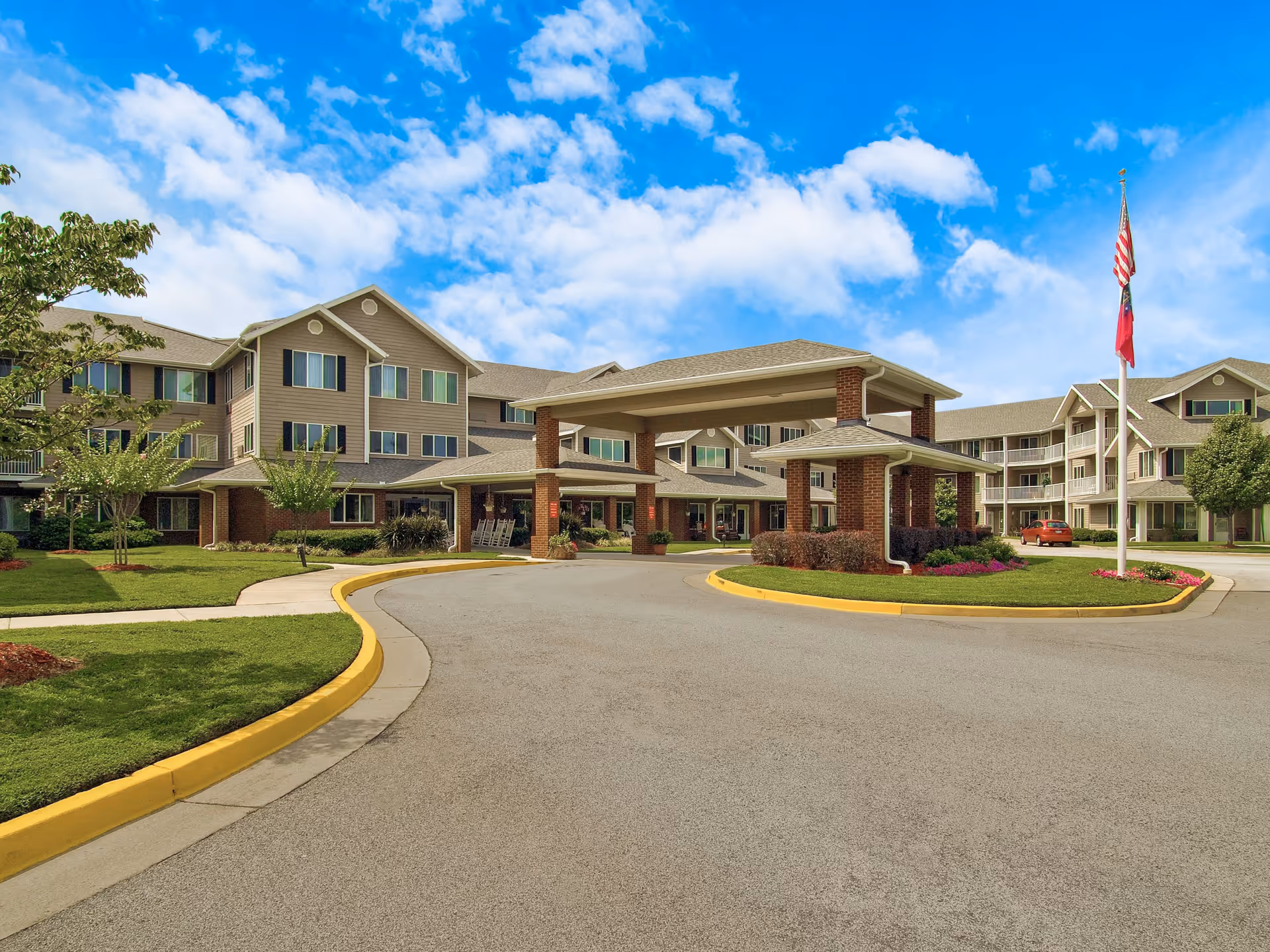 Exterior view of a large senior living facility building with multiple floors, a covered entrance supported by brick columns, well-maintained landscaping with green grass, trees, and flower beds, and an American flag on a flagpole under a partly cloudy blue sky.