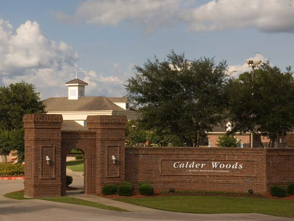 Entrance to Calder Woods, a Buckner Retirement Community, featuring a brick archway and brick wall with the community name, surrounded by green trees and landscaping under a partly cloudy sky.