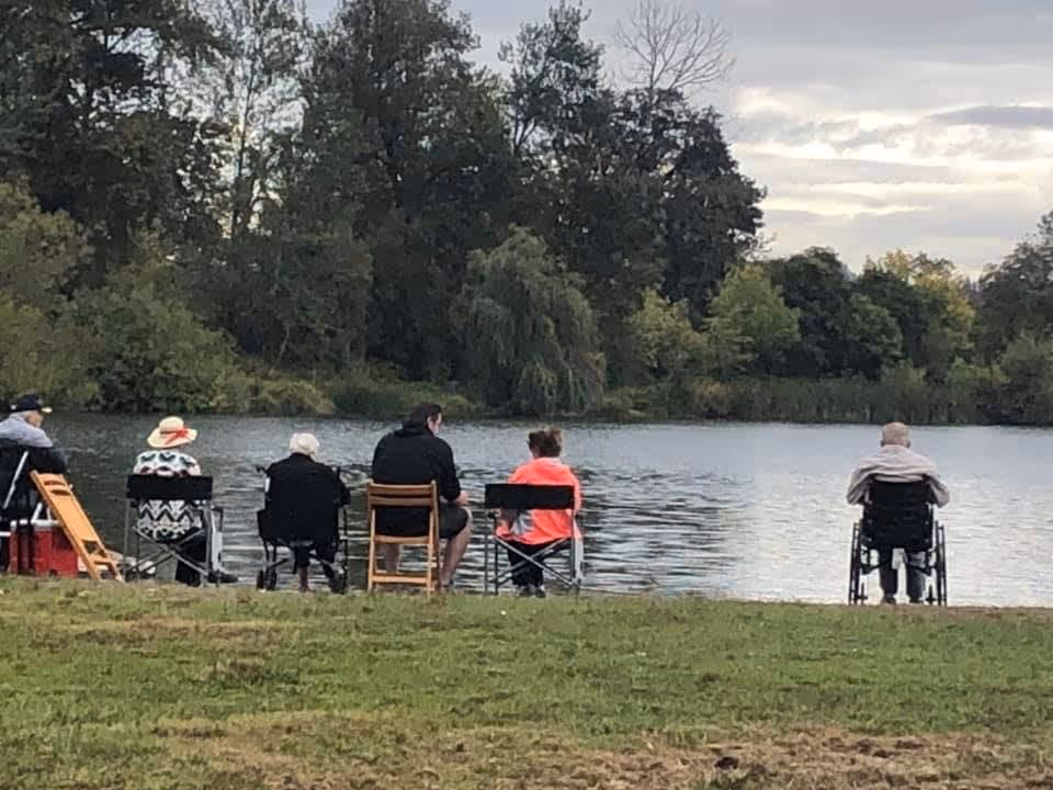 A group of six people sitting by a lake, facing the water. They are seated on various chairs and one person is in a wheelchair. Trees and bushes surround the lake under a cloudy sky.