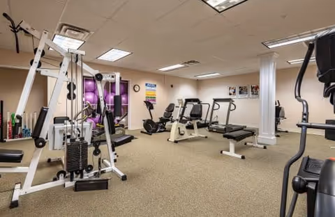 A fitness room with various exercise equipment including a multi-station weight machine, stationary bikes, treadmills, and exercise balls stacked in a corner. The room has beige carpet, white walls, and fluorescent ceiling lights.