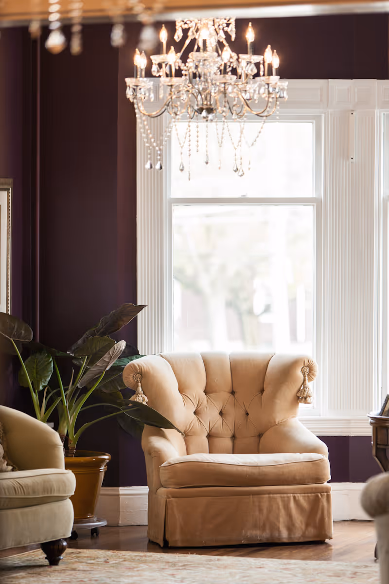 A cozy living room area with a beige tufted armchair featuring tassels on the armrests, a large window with white trim letting in natural light, a crystal chandelier hanging from the ceiling, a potted plant with large green leaves, and part of another beige armchair visible on the left side.