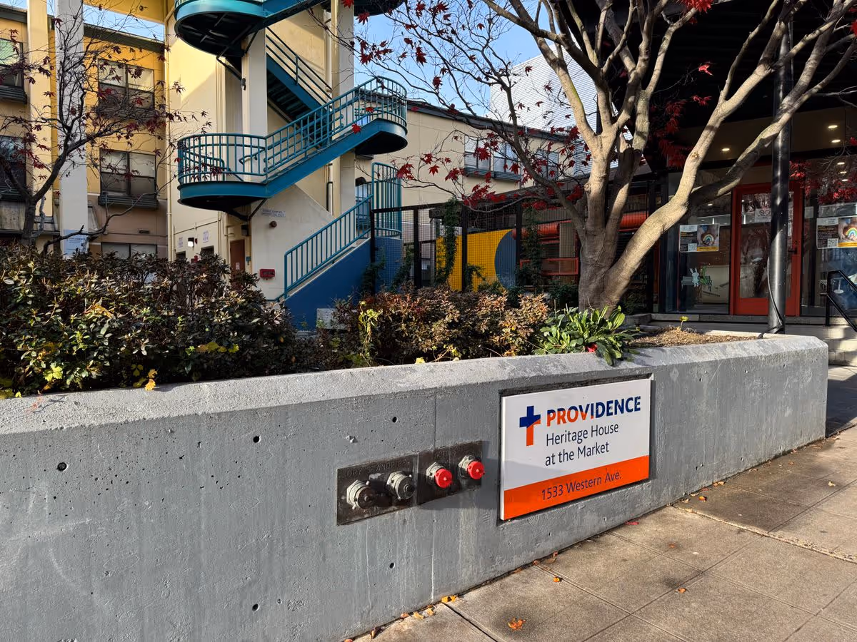 Exterior view of Providence Heritage House at the Market with the facility sign on a concrete planter, a tree, and a blue outdoor staircase by the entrance.