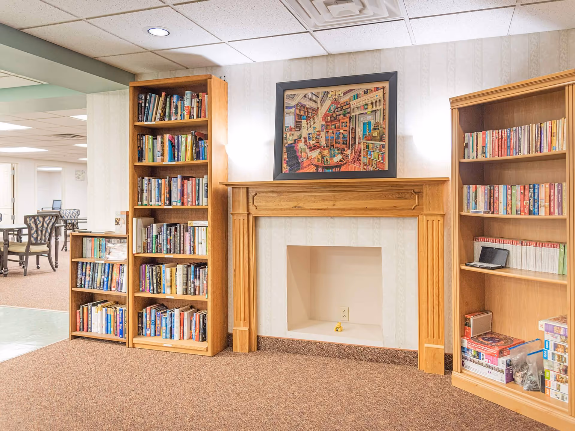 Interior view of a cozy reading area in a senior living facility featuring three wooden bookshelves filled with books, a wooden fireplace mantel with an empty hearth, and a framed picture of a colorful library scene hanging above the mantel. The room has carpeted flooring and a drop ceiling with recessed lighting. In the background, there are tables and chairs in an adjoining room.