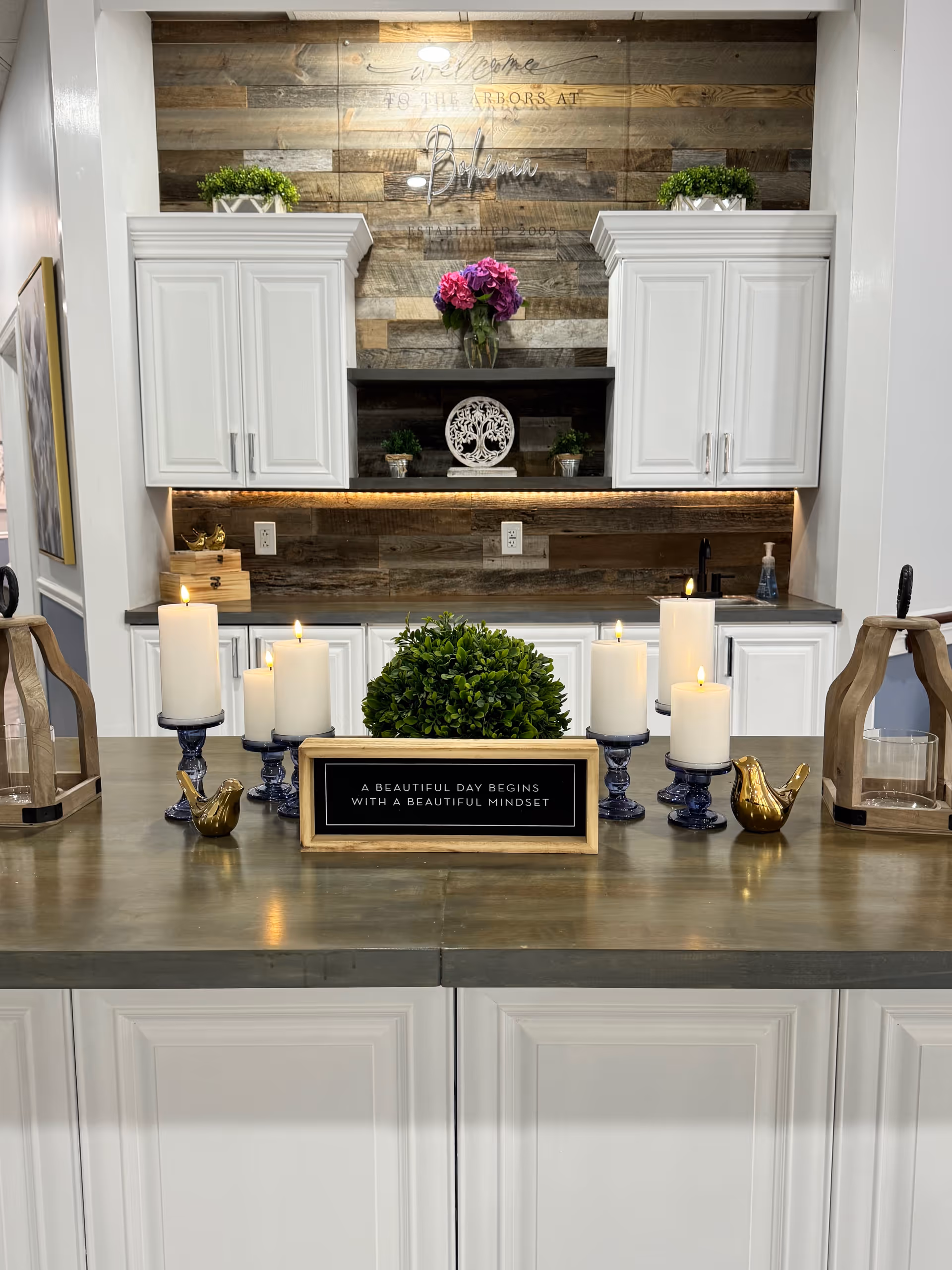A modern kitchen area with white cabinets and a wooden backsplash. On the countertop are several lit white candles on blue holders, a small green plant, and a framed sign that reads 'A BEAUTIFUL DAY BEGINS WITH A BEAUTIFUL MINDSET.' There are decorative items including two golden bird figurines and wooden lanterns on either side of the counter. Above the cabinets, a sign reads 'Welcome to The Arbors at Bohemia Established 2005.'