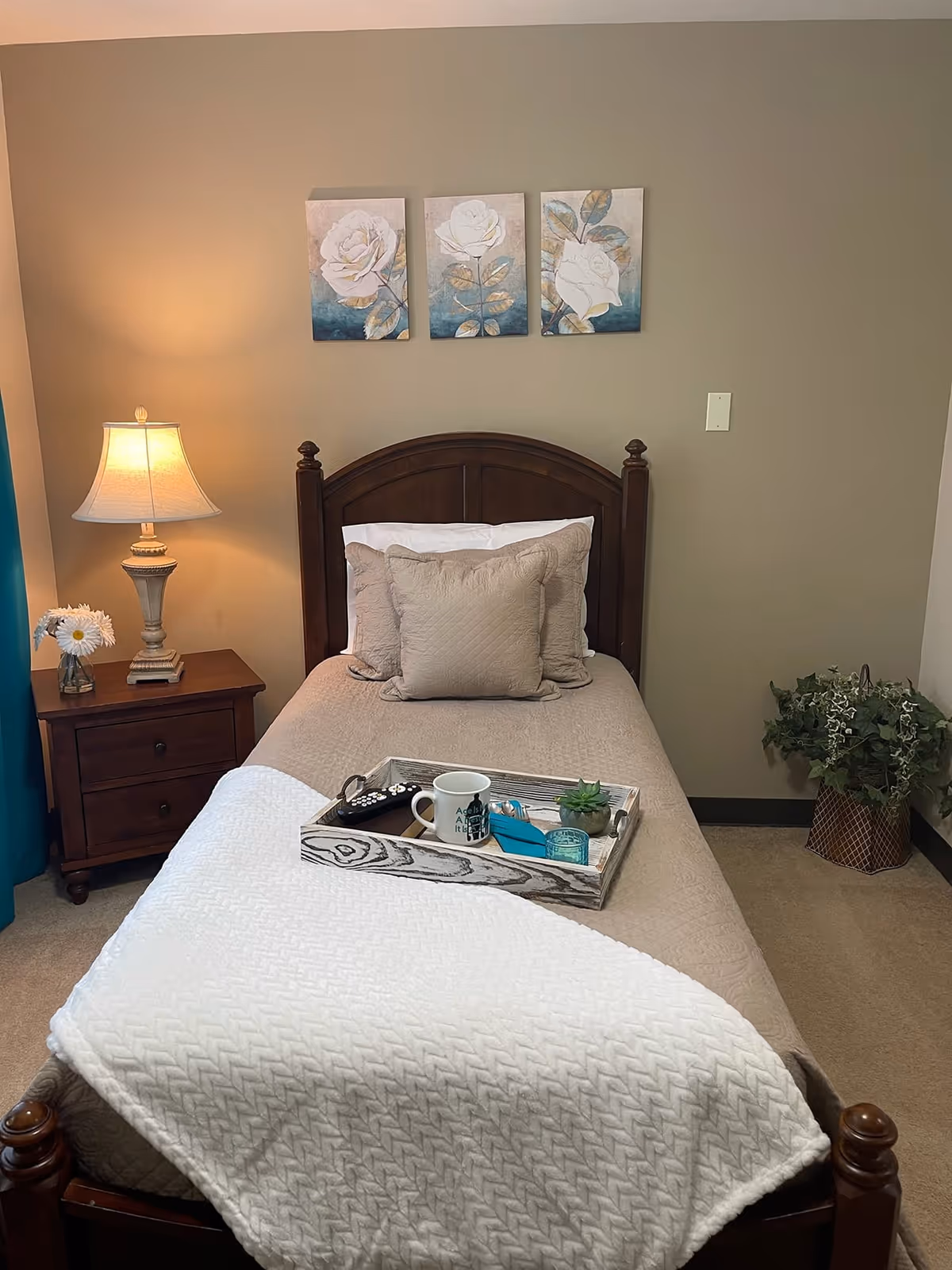 A neatly made single bed with a wooden headboard, decorative pillows and a tray, beside a nightstand with a lamp and wall art above.