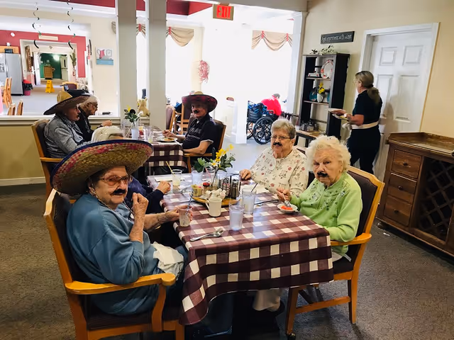 A group of elderly residents wearing sombreros and fake mustaches sit around a checkered table in a dining area, eating and socializing.