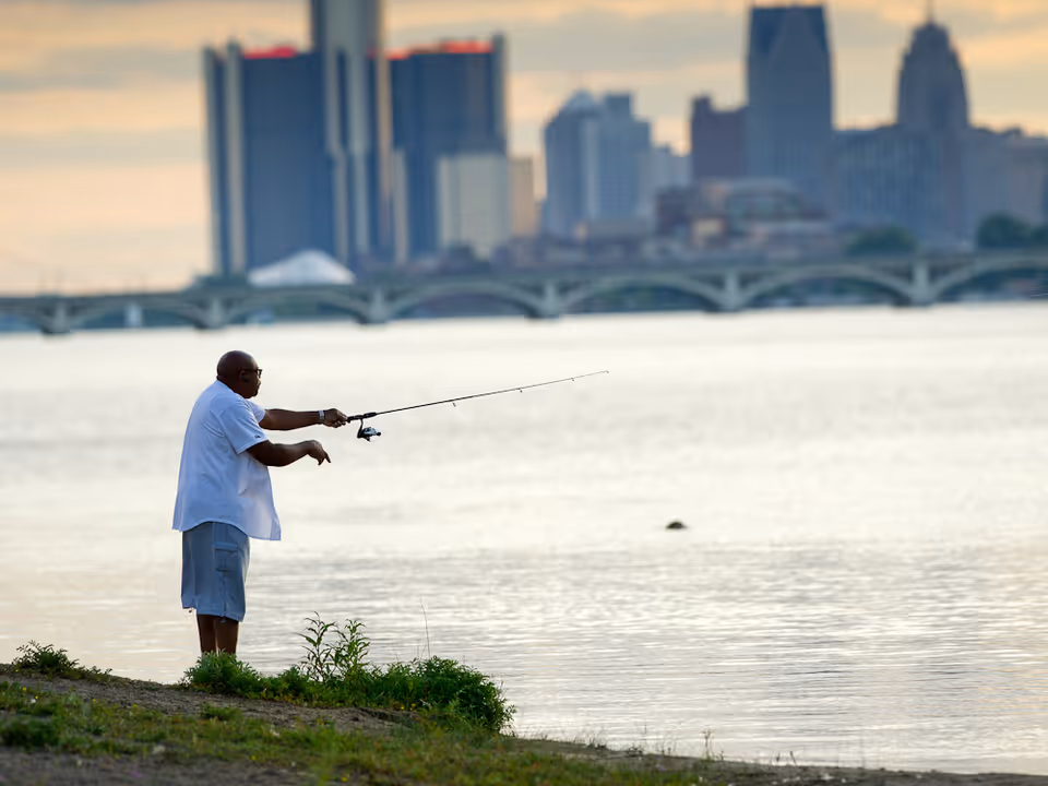 A person fishing from a grassy riverbank with a bridge and city skyline across the water in the background.