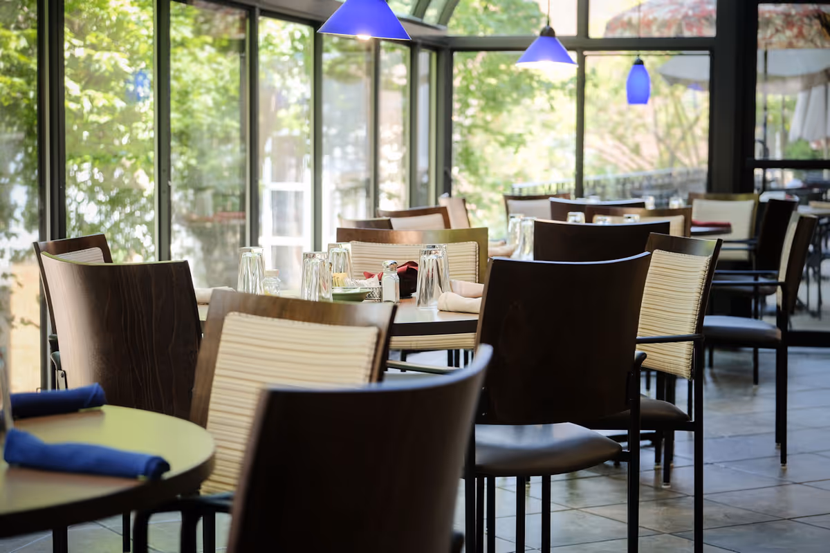A bright dining area with several tables and chairs arranged neatly. Tables are set with upside-down glasses, napkins, and condiments. Large windows allow natural light to fill the space, and blue pendant lights hang from the ceiling.