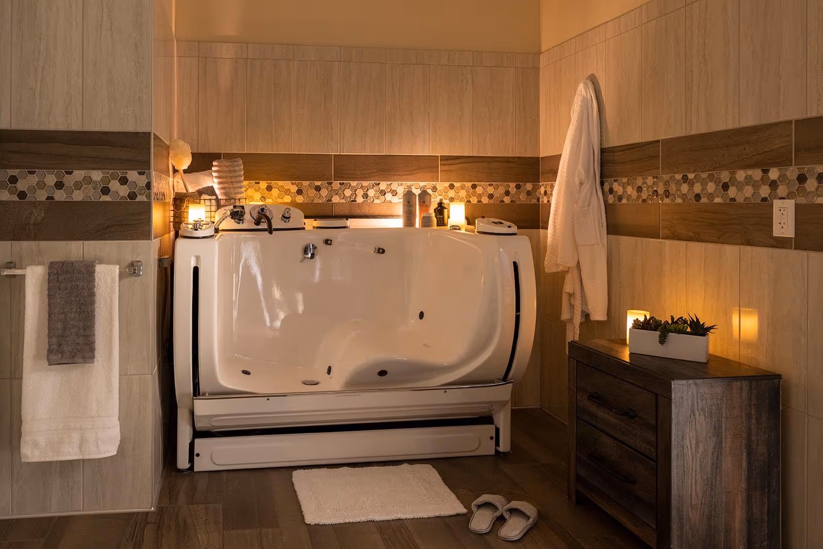 A warm, softly lit bathroom featuring a walk-in bathtub with a door, surrounded by beige and brown tiled walls with a decorative mosaic strip. A white bathrobe hangs on the wall, and a wooden cabinet with a small plant and candle is placed nearby. Towels hang on a rack to the left, and slippers and a bath mat are on the floor in front of the tub.