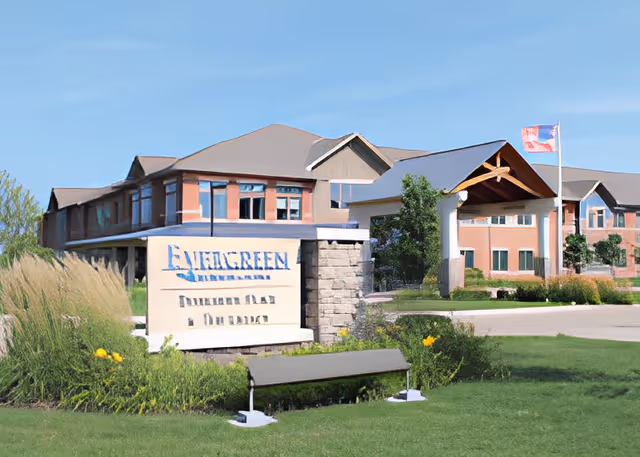 Front exterior of the Evergreen senior living facility showing the entrance canopy, sign, landscaped lawn, and flag.