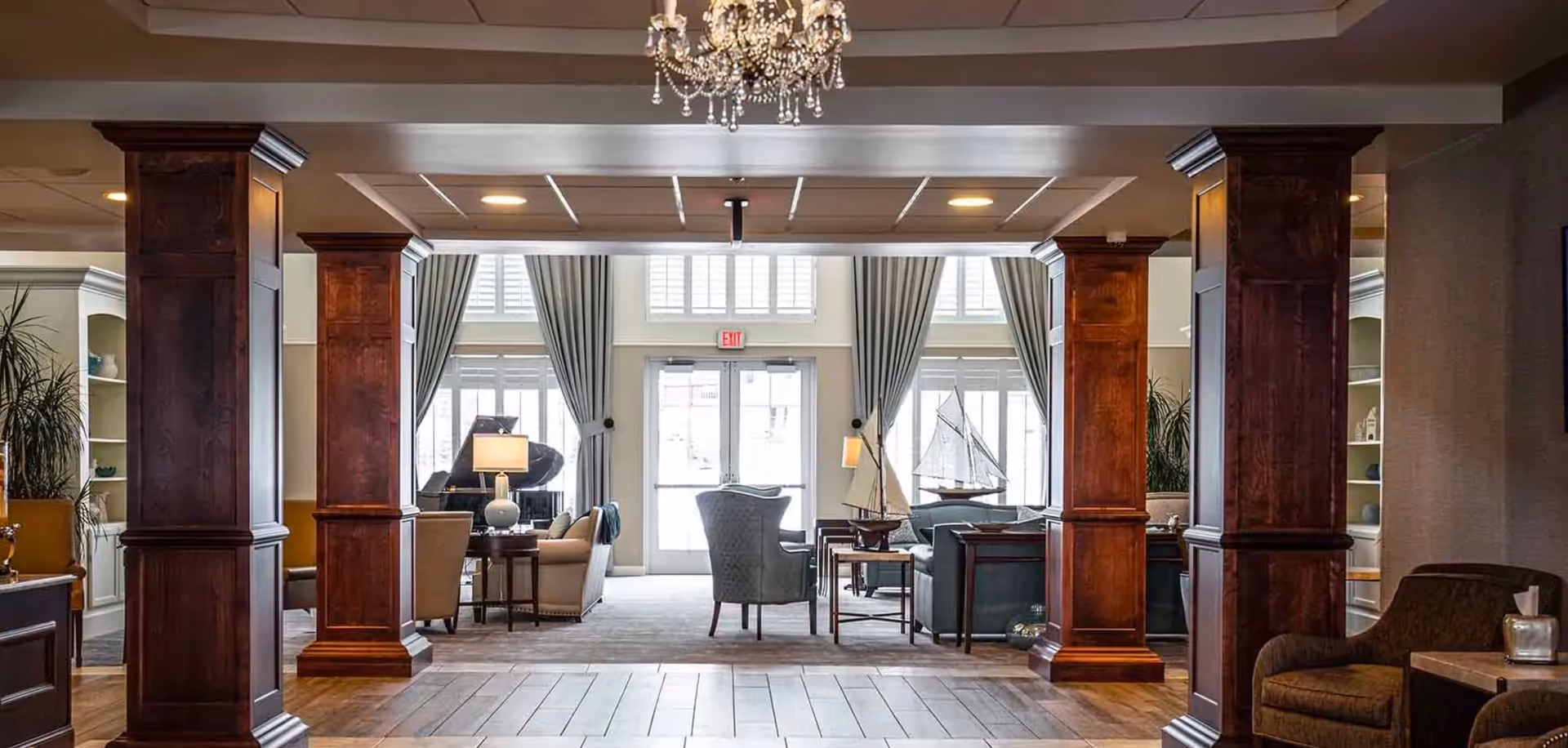 Spacious elegant lobby/lounge with wooden columns, chandelier, sofas and chairs arranged facing tall windows and glass doors.